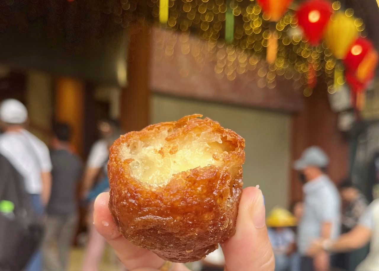 Food in Hanoi: The Best Old Quarter Restaurants 4 A Vietnamese donut fried with honey and sesame seeds is being held with two fingers in front of blurred red and yellow lanterns on the street in Hanoi.