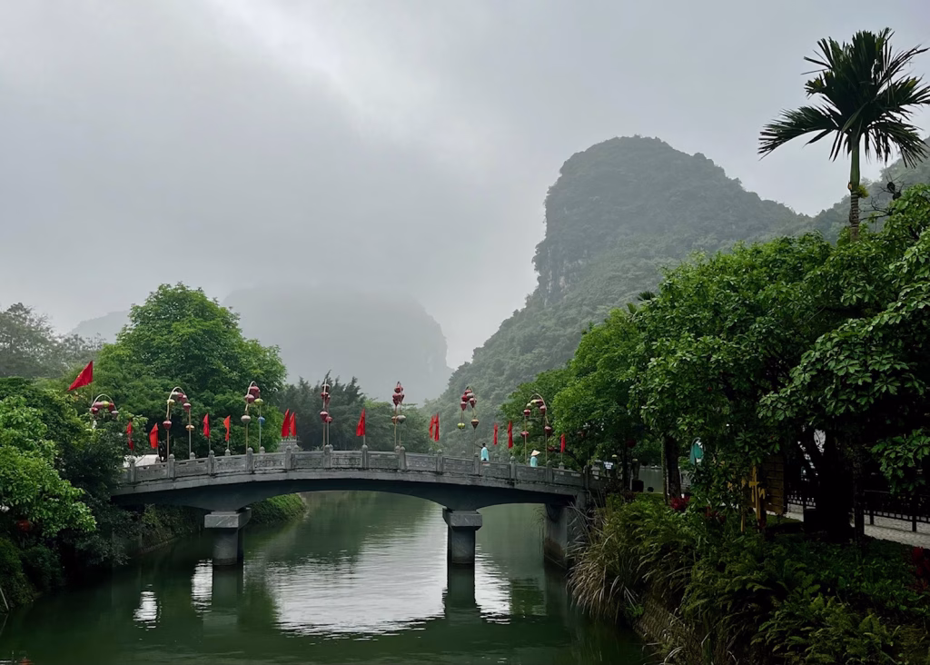 Tam Coc Bicycle Tour: The Perfect Self-Guided Route 10 The misty entrance to the Trang An boat complex. There is a bridge with bright red Vietnam flags in the distance.