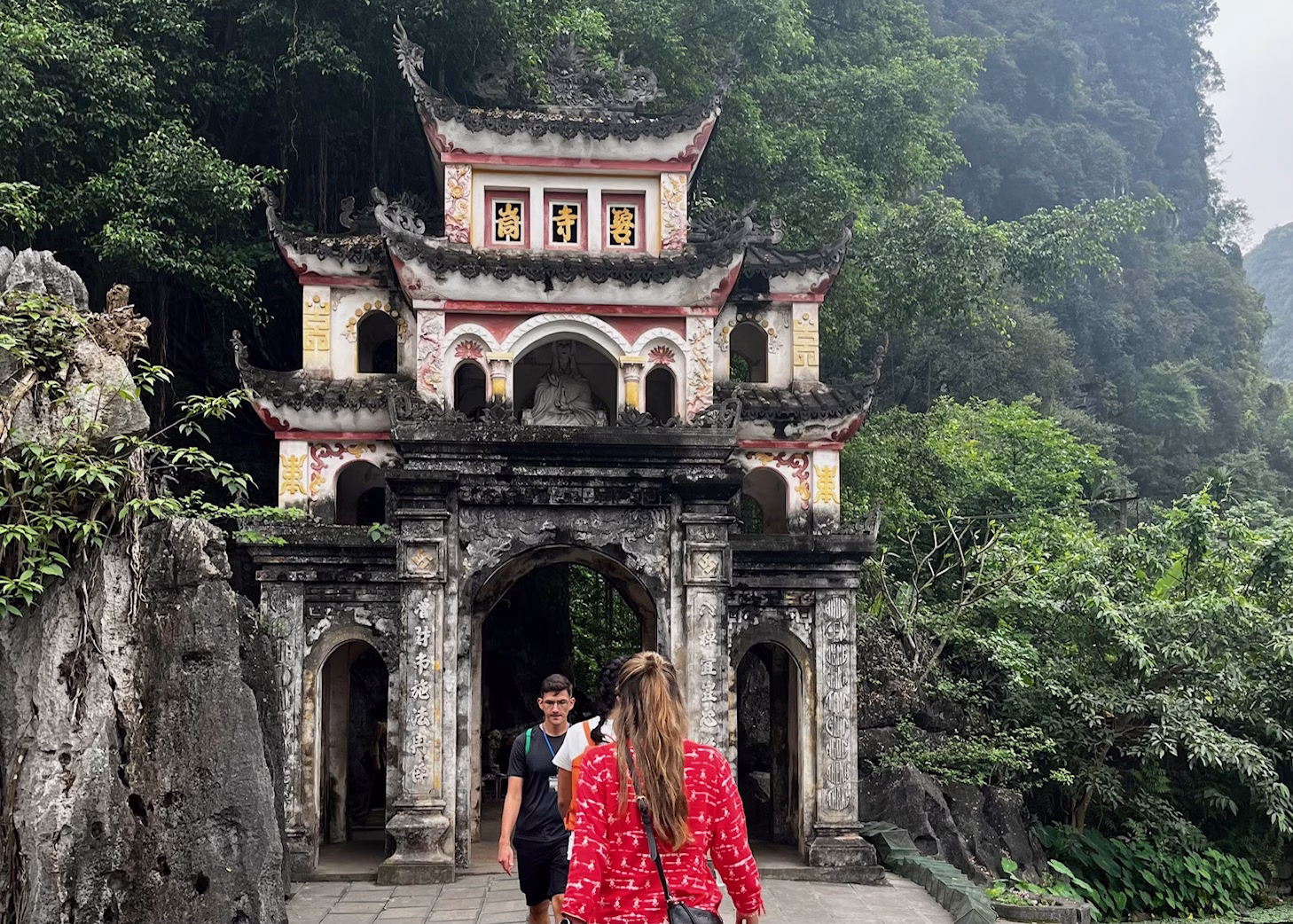 Tam Coc Bicycle Tour: The Perfect Self-Guided Route 6 People walking along the path over the river leading to Bich Dong Pagoda are standing in front of the entrance. There is a green mountain behind the pagoda and mountains on the side of the bridge.