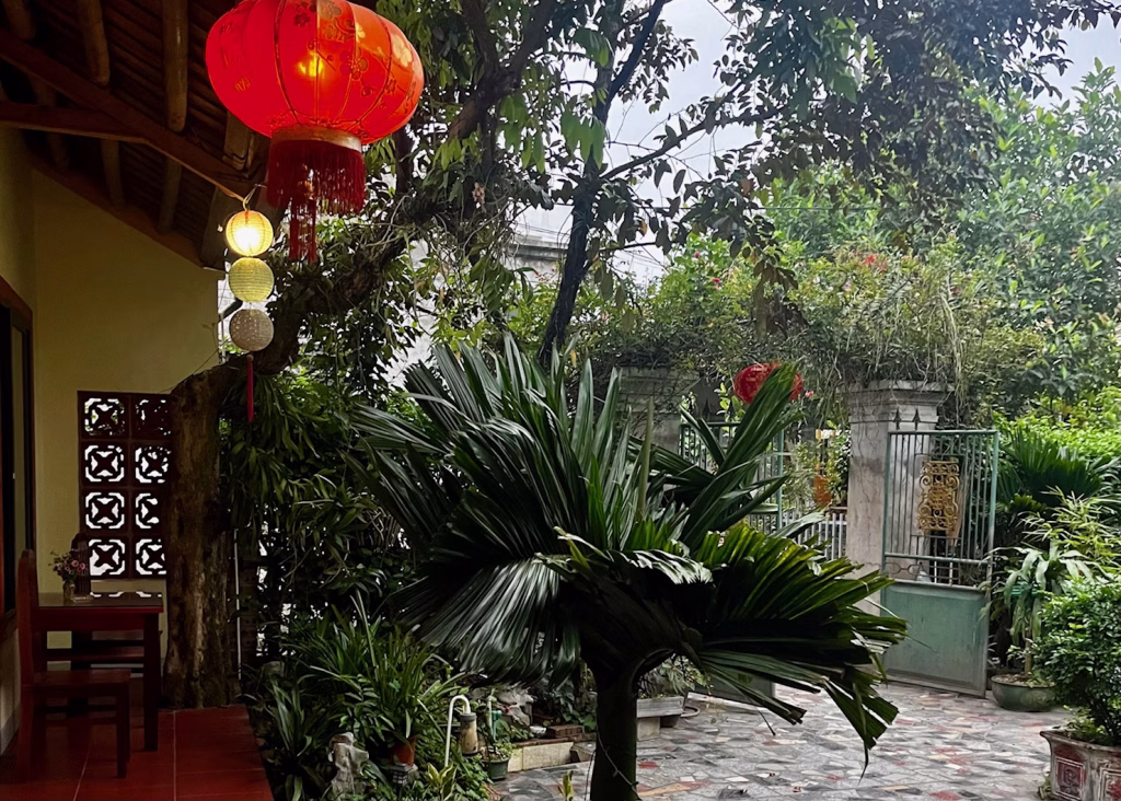 Tam Coc Bicycle Tour: The Perfect Self-Guided Route 9 The green garden of the homestay called Tam Coc Lotus Flower Homestay. There are red lanterns hanging from the ceiling and wooden tables and chairs sitting on the porch.