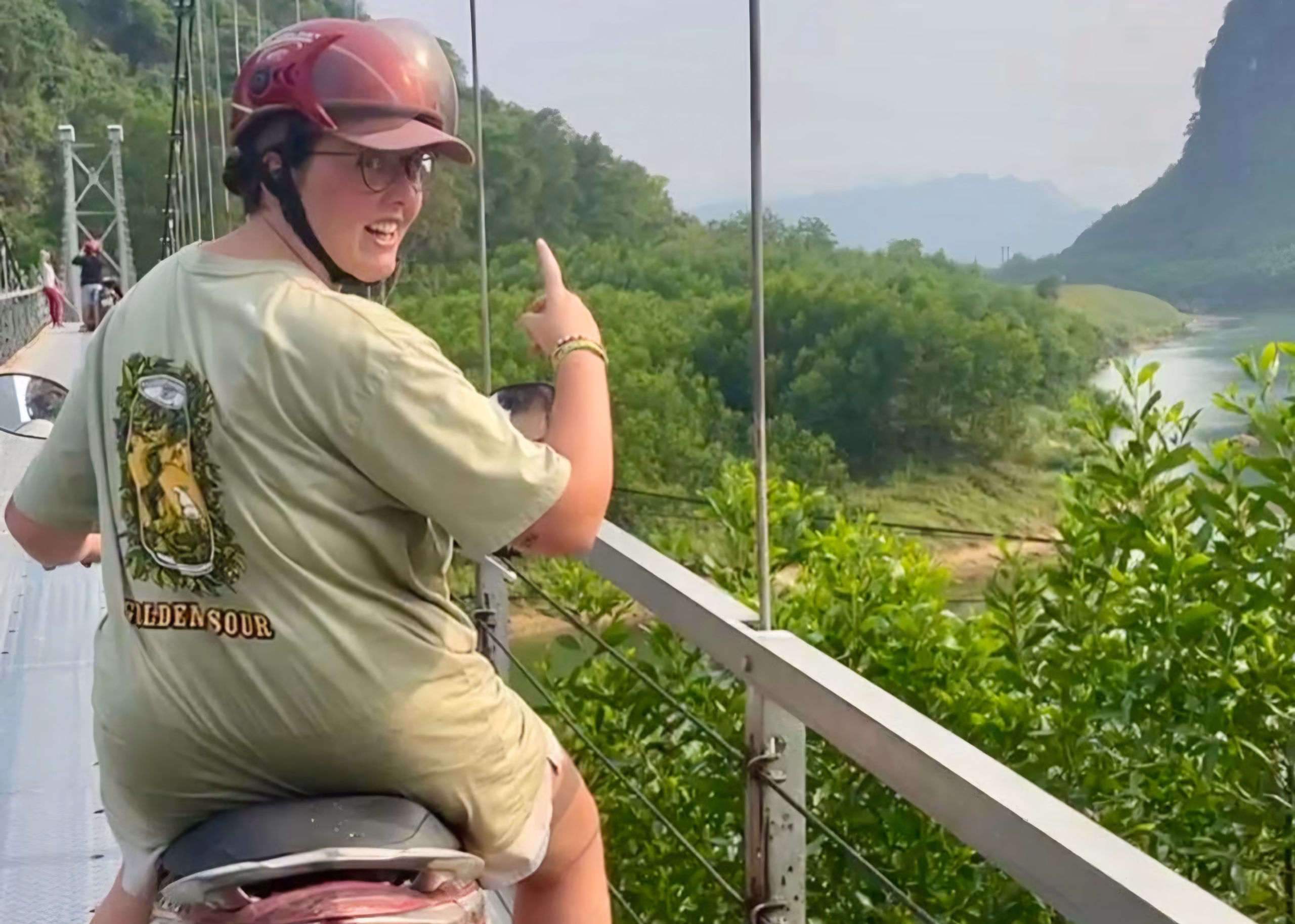 12 Best Things to Do in Phong Nha (Caves, Countryside & Motorbike Adventures) 10 A girl in a green t-shirt is sat on a motorbike on a bridge in Phong Nha. She is pointing to the green scenery and river below