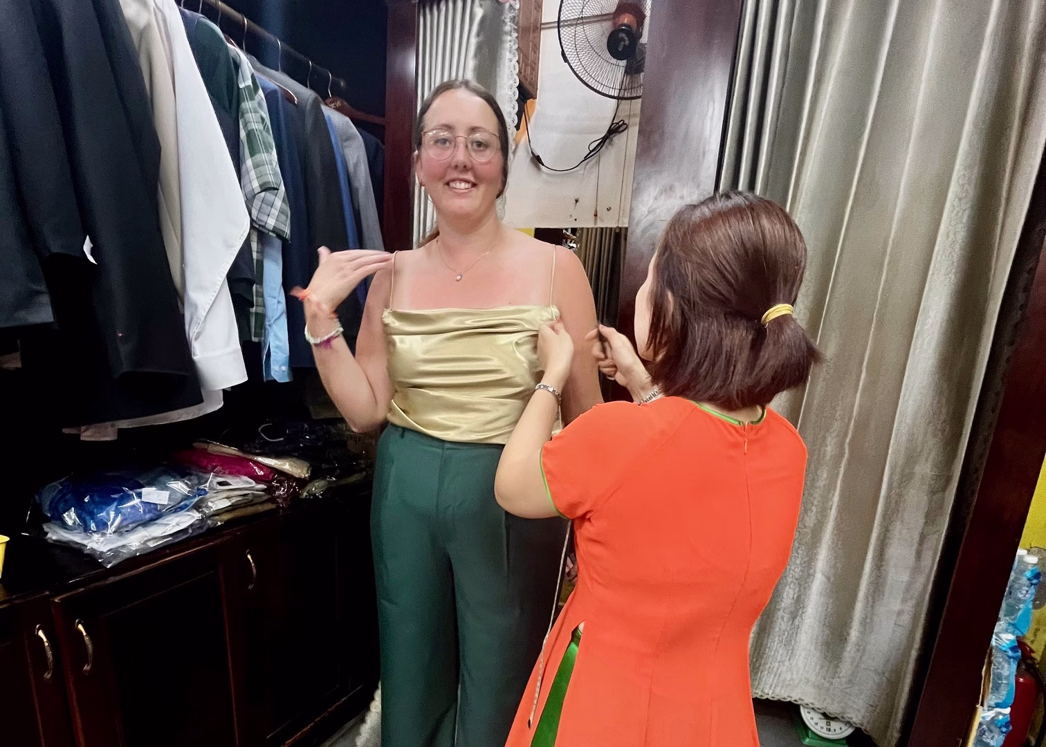 Hoi An Itinerary: 3 Days of Food & Lantern-Lit Charm 2 A girl is having clothes tailored during a 3 day Hoi An itinerary. She is wearing a gold top and green trousers, and the seamstress is adjusting the straps on the top