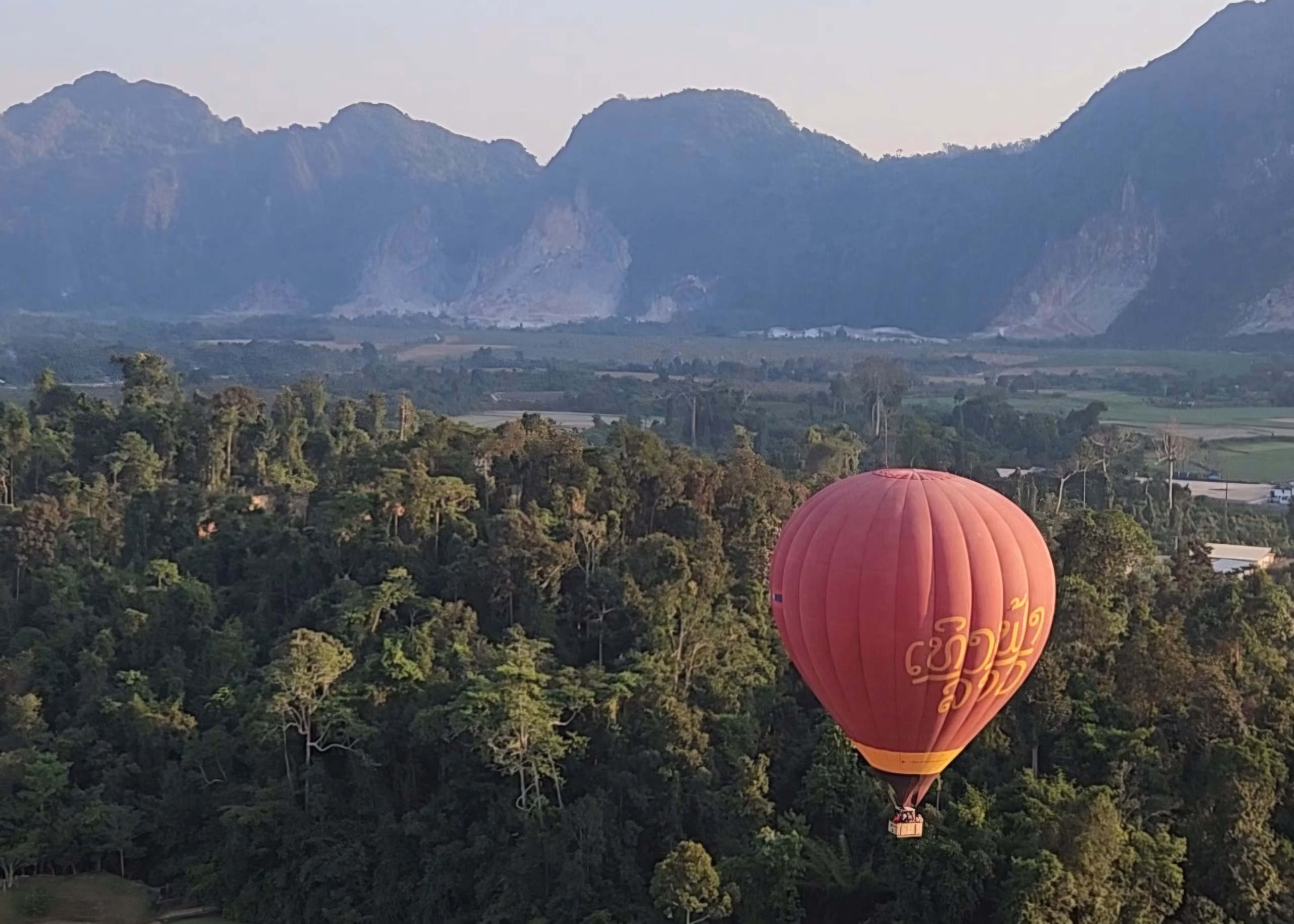 A closer view of another balloon taken from the basket of a flying hot air balloon. You can see the trees and mountains of Vang Vieng below.