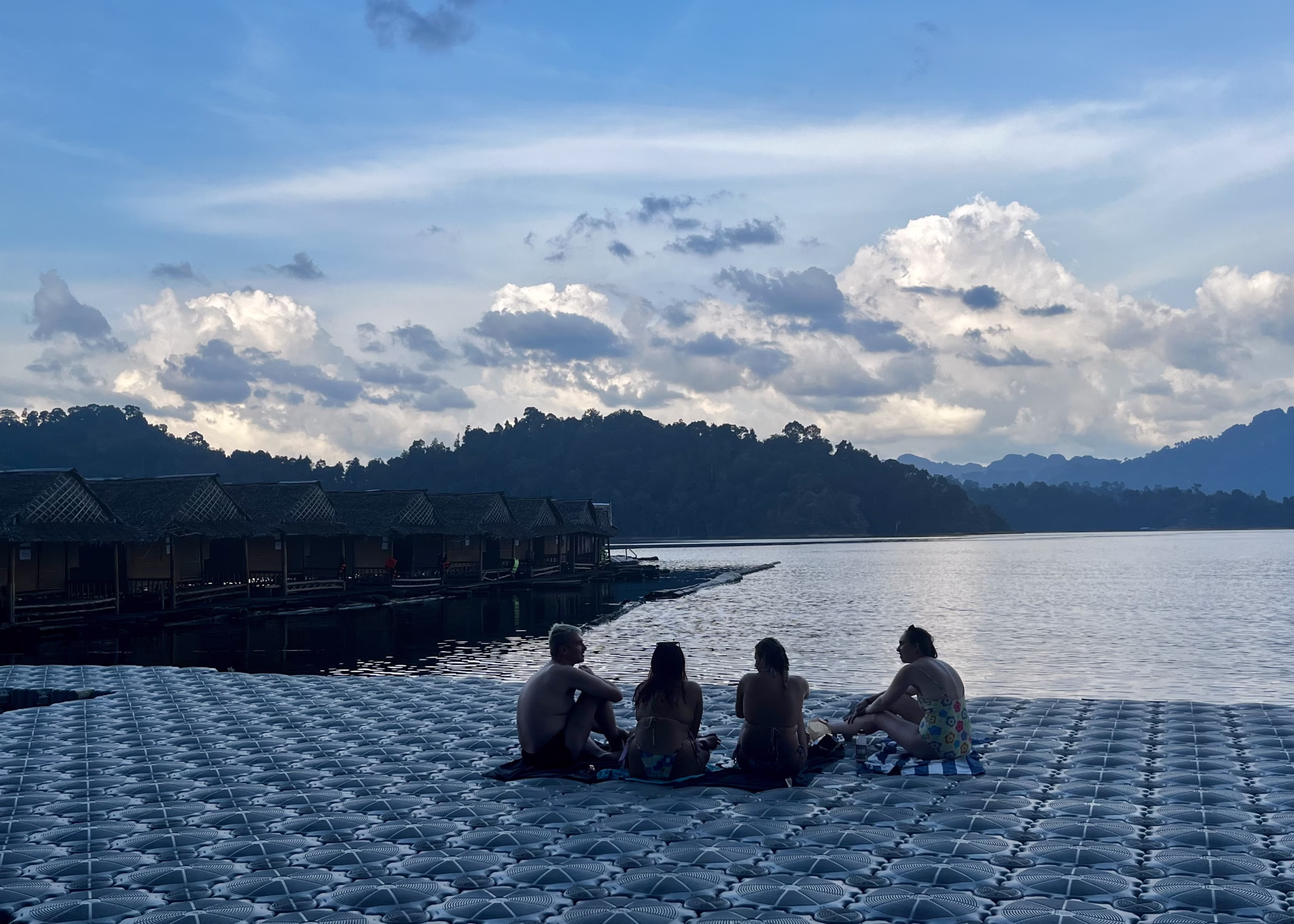 Khao Sok National Park Floating Bungalows: What to Expect 6 A group of 4 people are sitting on the plastic dock at the Khao Sok National Park Floating Bungalows. The sun is beginning to go down, so the people are covered in shadows and the mountains on the lake are a little hazy.