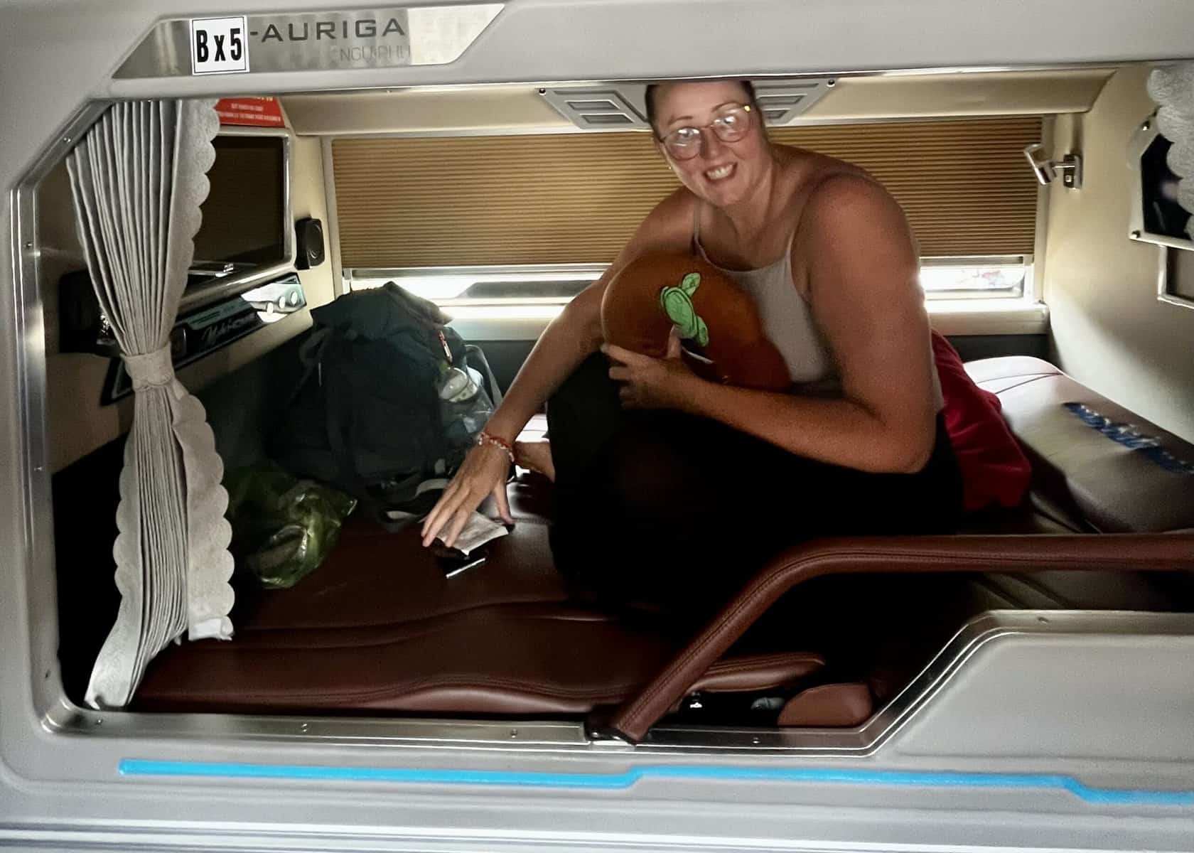 Where to Stay in Hoi An (3 Best Areas + Stays We’d Book) 11 A tall woman is sitting up in her top bunk on a sleeper bus in Vietnam. She is holding her neck pillow and smiling.