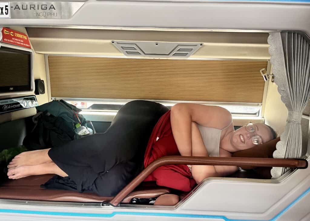 Laos or Vietnam: How to Choose the Best Country for Your Trip 19 A tall woman is laying down in her top bunk on a sleeper bus in Vietnam. She is holding her neck pillow and smiling. Her legs are bent as she doesn't fit fully in the built-in bed.