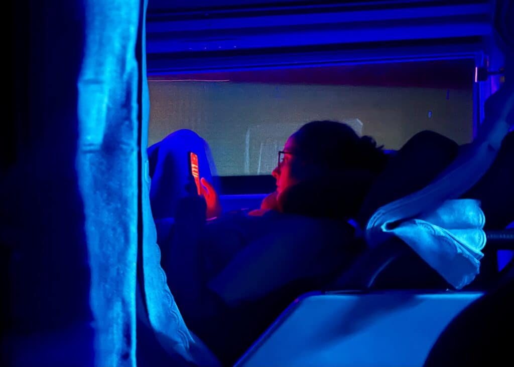 Bangkok to Chiang Mai Travel Guide — How to Get There by Train, Bus & Plane 7 An outline of a woman in her bed on a sleeper bus in Vietnam. The blue light from the bus is glowing all around her and the red light from her phone is lighting up her face.