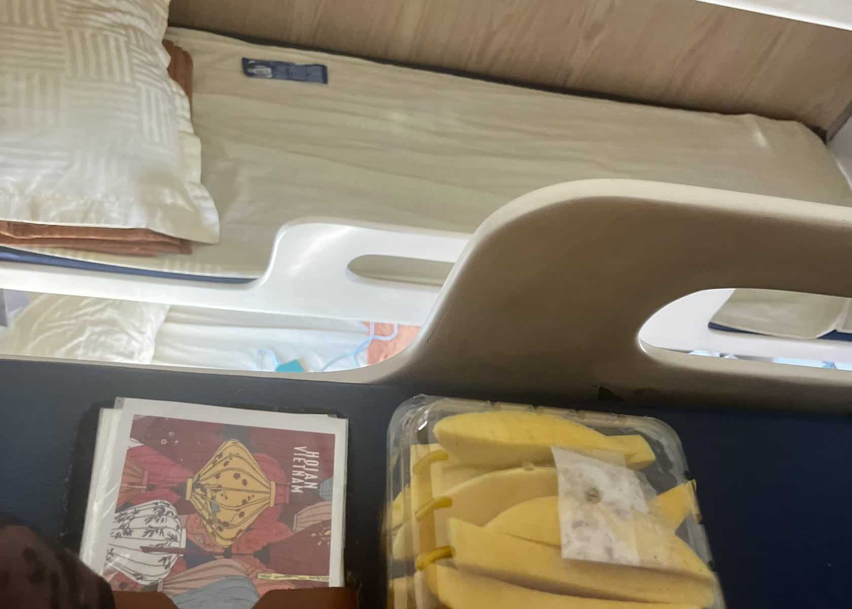 Overnight Sleeper Train in Vietnam: A Guide for First-Timers 3 Mangos in a plastic container and a kindle are sitting on the top bunk of an overnight sleeper train in vietnam. You can see the lower beds on the opposite side.