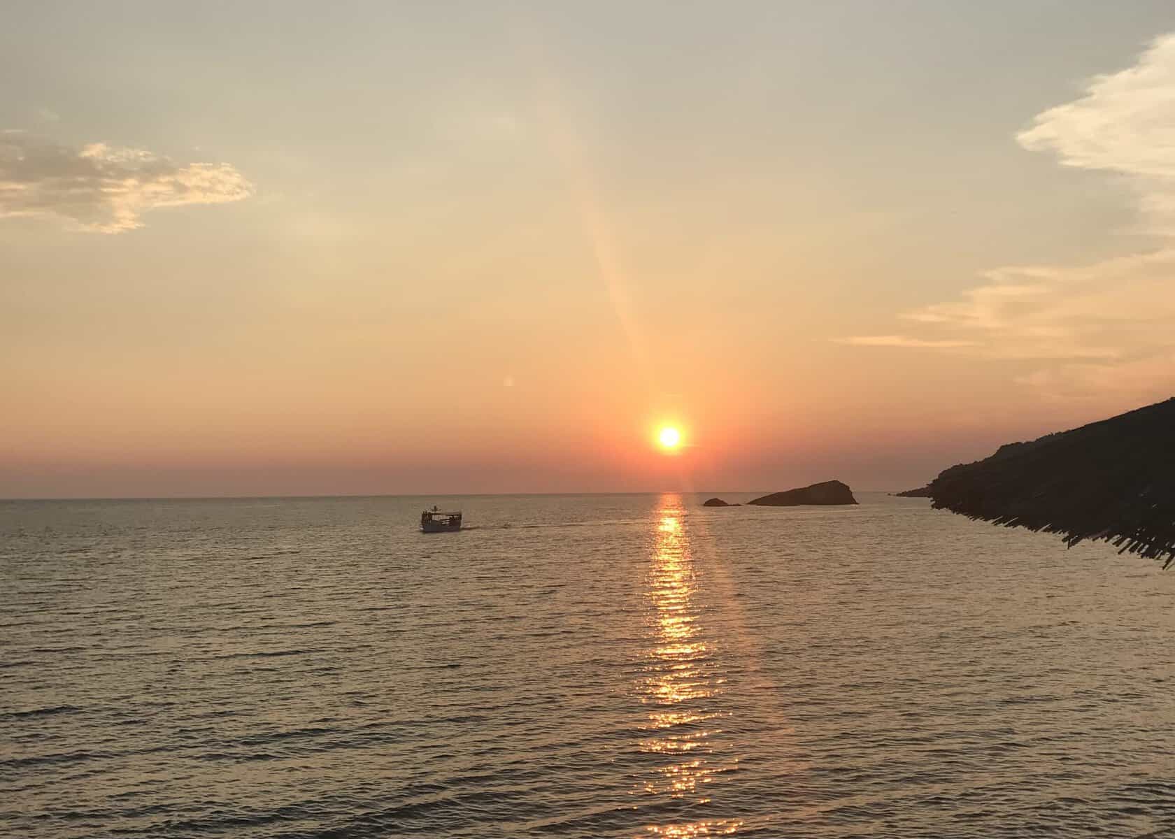 One Week South Albania Itinerary: Beaches, Castles & Culture 15 the sun is setting over the water as seen from a cliff in Himarë. The sun is reflecting orange and pink over the water.