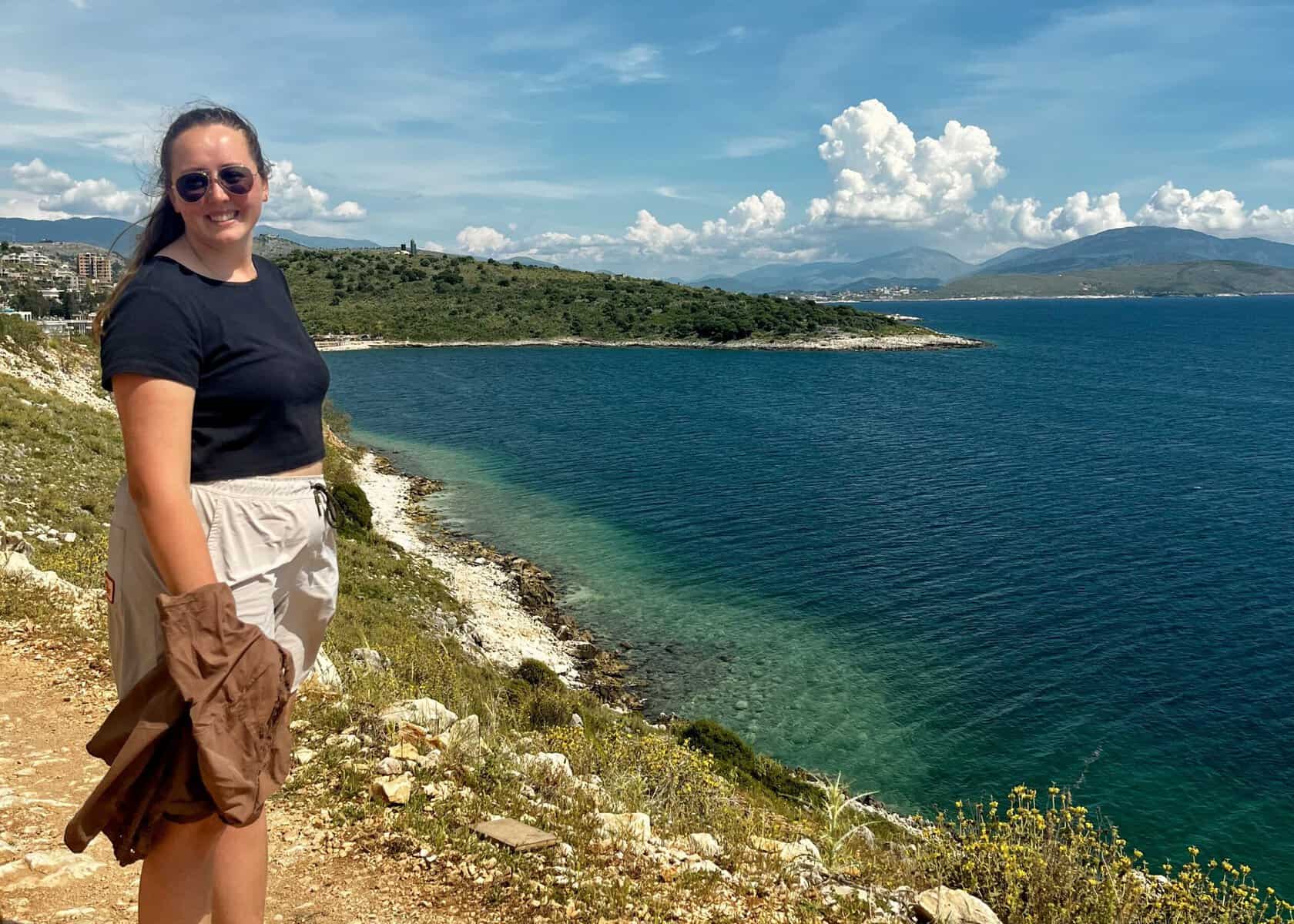 One Week South Albania Itinerary: Beaches, Castles & Culture 5 A woman is standing on the cliff above bright blue water in while walking to one of the beaches in Himarë.