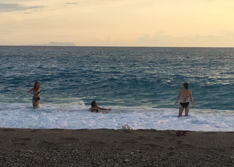 Three women are splashing in the water at Himarë Beach at dusk.
