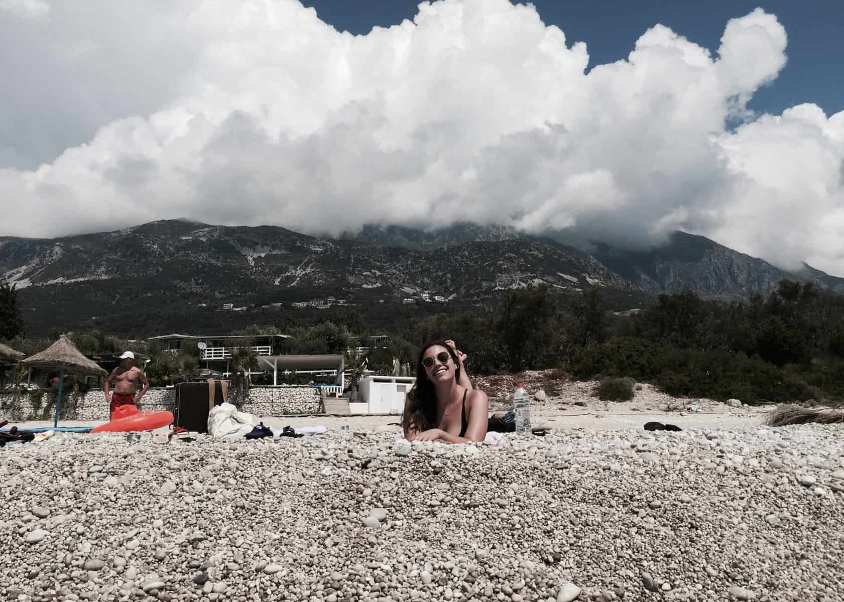 One Week South Albania Itinerary: Beaches, Castles & Culture 4 A woman is smiling and laying on a the beach in Dhermi, Albania. The beach here is rocky and there are mountains and white fluffy clouds behind her.