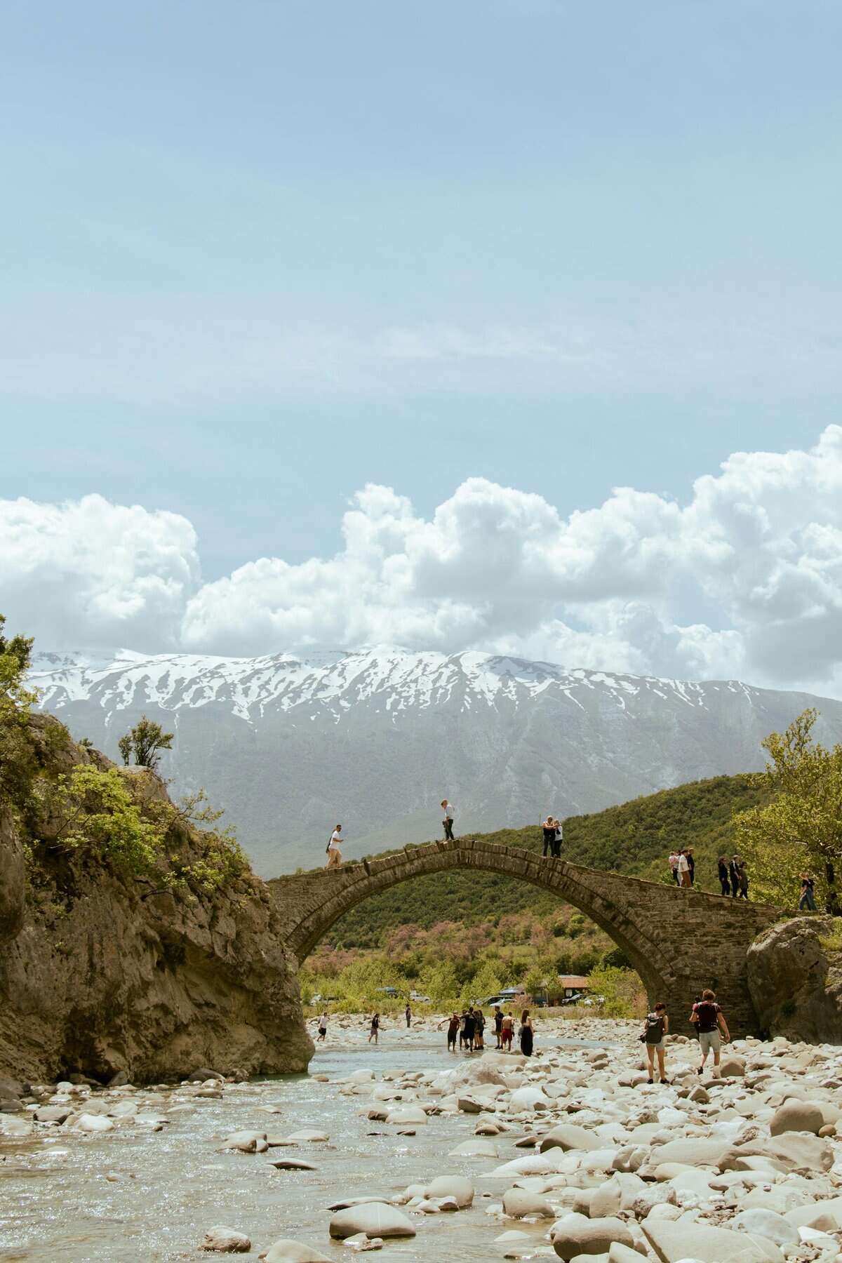 One Week South Albania Itinerary: Beaches, Castles & Culture 10 Scenic view of a historic stone bridge over a river in Përmet, Albania, featuring people and mountain backdrop.
