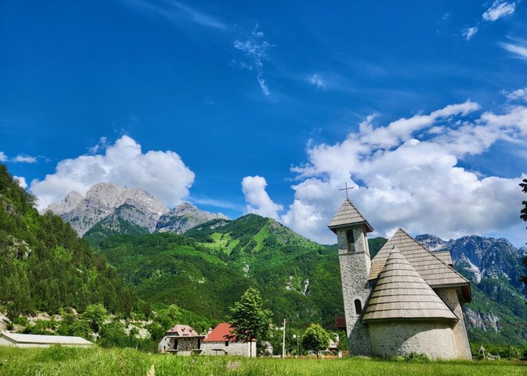 An old church in the village of Theth, Albania is sitting in front of a blue sky and green mountains.
