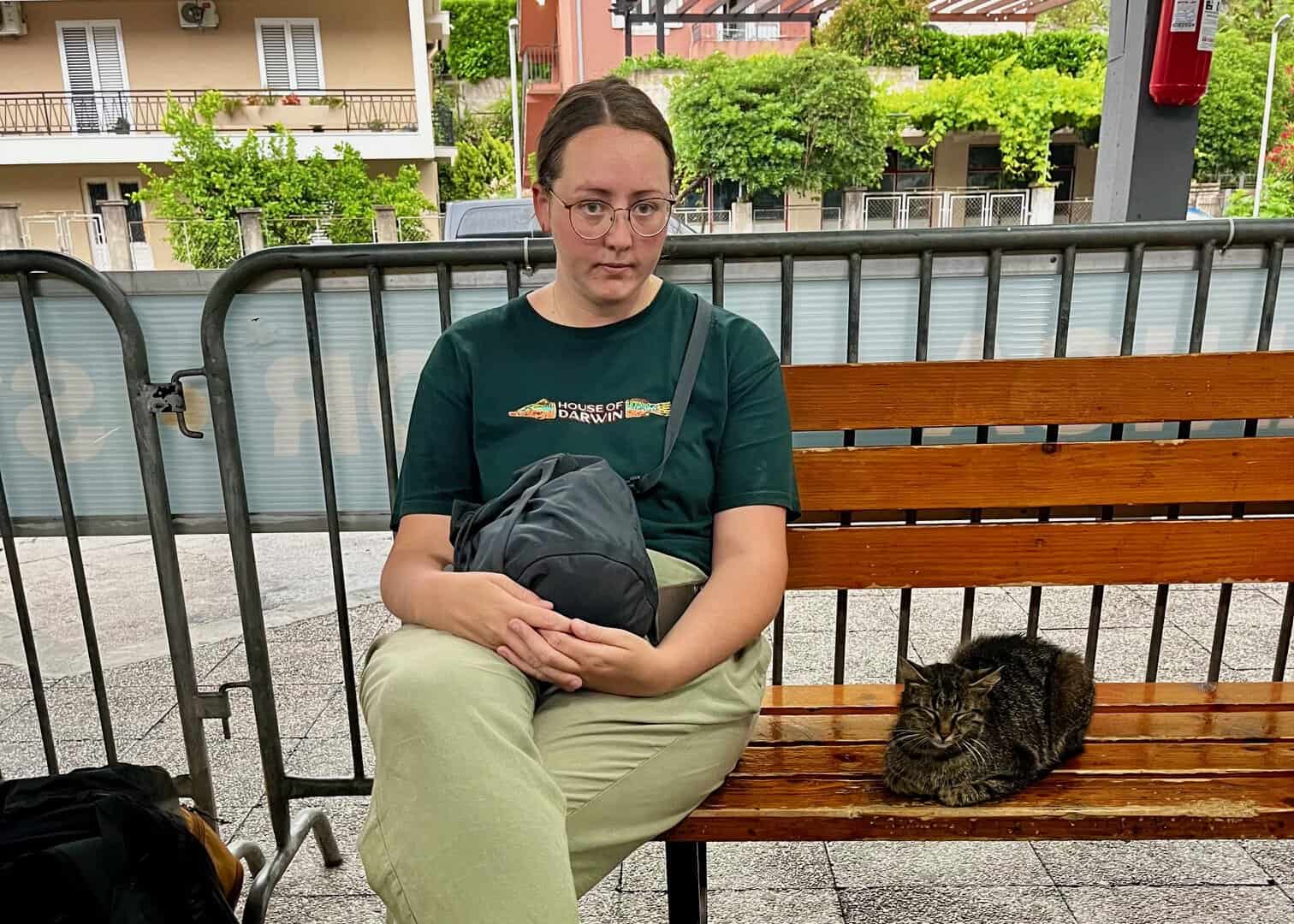 A woman in a green tshirt and pants is waiting on a wooden bench at a bus station in Albania while waiting on a bus to leave. There is a cat sitting next to her.