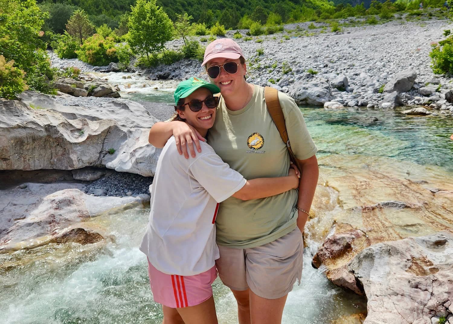 Where to Stay in Theth: Best Value Guesthouses 5 Two women in hiking attire are hugging and smiling in front of a blue river in Theth, Albania.