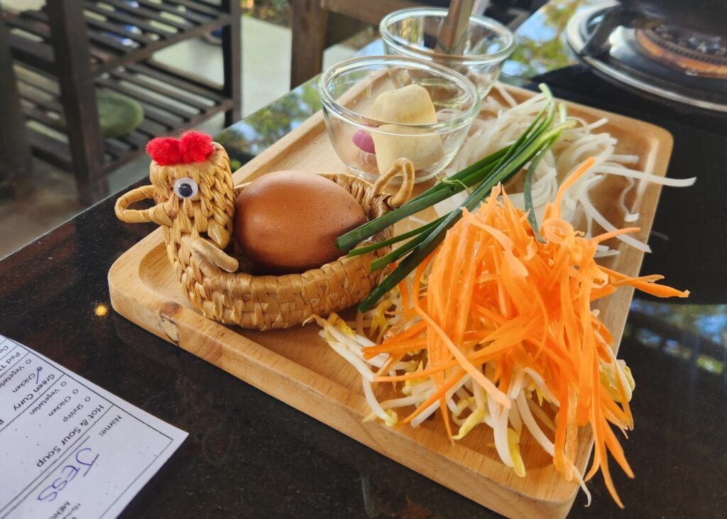 Our Honest Grandma’s Home Cooking School Review 2 Ingredients to begin a cooking class at Grandma's Home Cooking School in Chiang Mai are sitting on a wooden board. There is an egg in a chicken-shaped wicker basket, as well as ginger, carrots and green onions on the board.