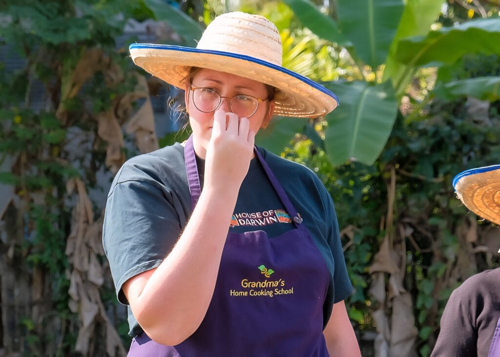 Our Honest Grandma’s Home Cooking School Review 6 A woman in a navy blue apron and brown sun hat is smelling herbs from the garden an Grandma's Home Cooking School in Chiang Mai.