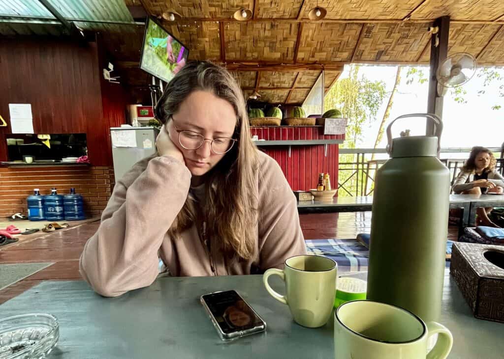Laos or Vietnam: How to Choose the Best Country for Your Trip 28 Young woman with glasses resting her head on hand at a table in a tropical, cozy cafe setting.