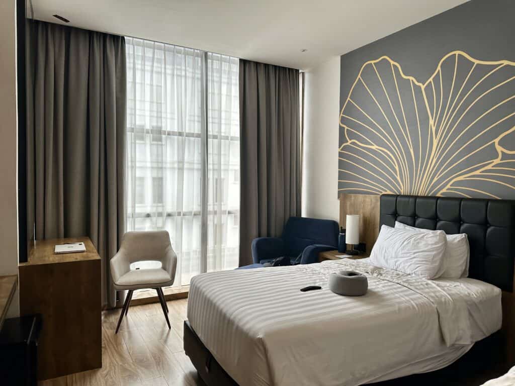 A hotel in Medan, Sumatra with grey walls and curtains, wood floors, and a neatly made bed with white sheets. A hotel upgrade is a great idea when buying holiday gifts for travelers