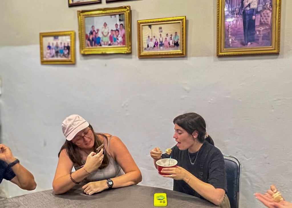 Bangkok Food Tour: Our Honest Review of Chinatown’s Best Street Eats 6 Two women are sitting at a table in a small shop on a Bangkok food tour. One of them is making a funny face while trying a shrimp dumpling and the other is taking a photo of her with her phone. Behind the table is a series of photos showing the Thai Princess eating at the restaurant.