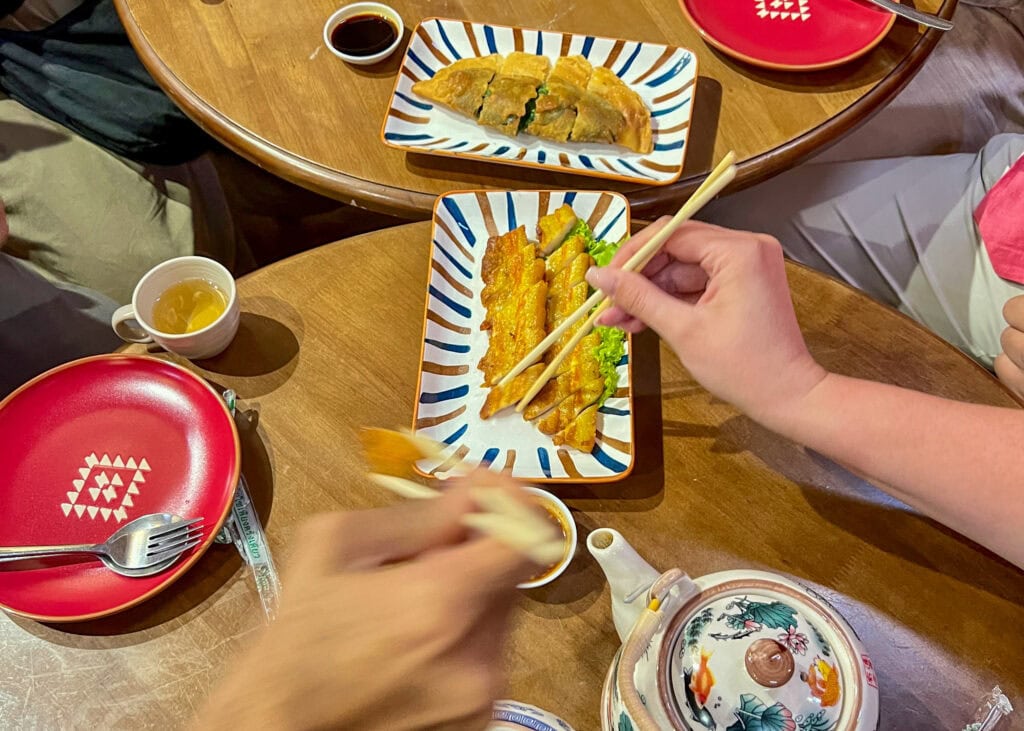 Bangkok Food Tour: Our Honest Review of Chinatown’s Best Street Eats 8 An overhead photo of chive pancakes and crispy pork served at a dim sum restaurant on a Bangkok food tour. Two different people are holding chopsticks about the food waiting to pick it up.