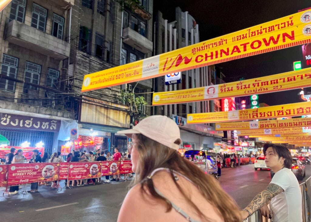 Bangkok Food Tour: Our Honest Review of Chinatown’s Best Street Eats 11 A woman is leaning on a railing in the Bangkok Chinatown night market and looking out over the road. Above her there are many yellow signs in Thai and Manderine advertising the vegetarian festival.