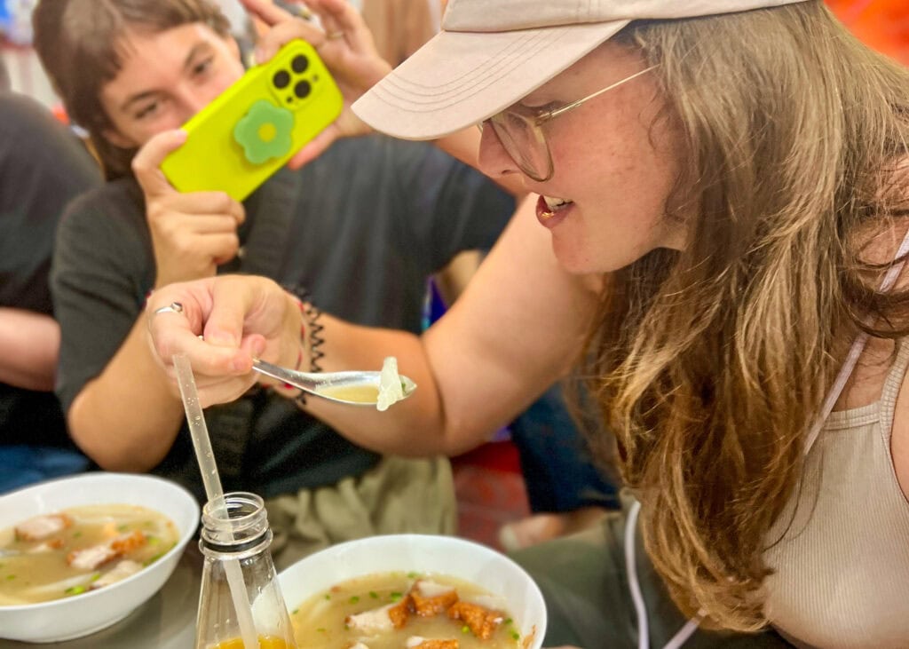 Where to Go in Thailand for the First Time: A Budget Traveler’s Guide 6 A woman in a pink hat is holding a soup soup with a noodle on it ready to take a bite from a dish served on a Bangkok food tour. Someone with a bright green phone is taking her photo in the background.
