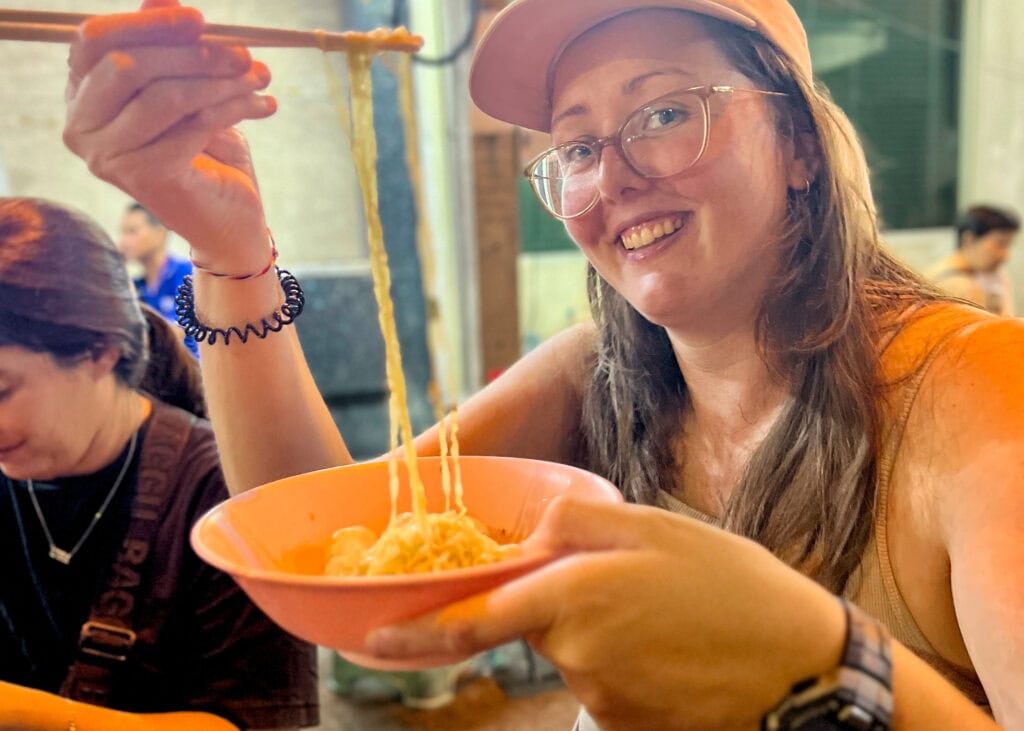 Bangkok Food Tour: Our Honest Review of Chinatown’s Best Street Eats 16 A woman is holding up noodles over a bowl of fish ball soup with chopsticks.