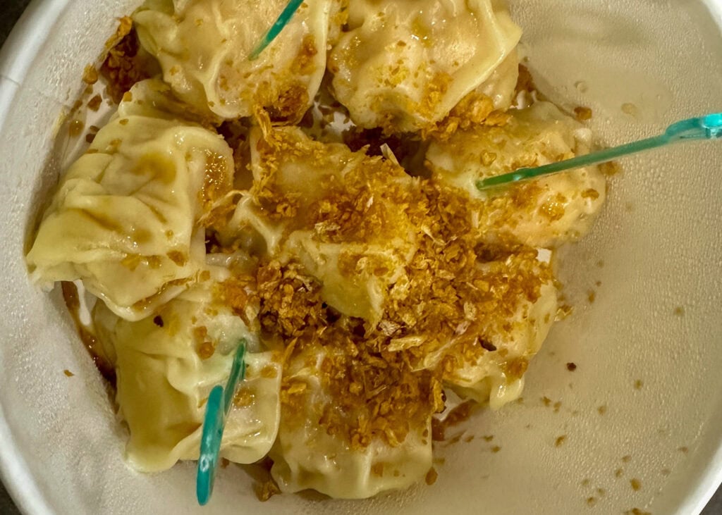 Bangkok Food Tour: Our Honest Review of Chinatown’s Best Street Eats 7 A close up of shrimp dumplings in a paper bowl covered with a ton of dried garlic and sour fish sauce. There are two blue plastic forks stuck into the dumplings.
