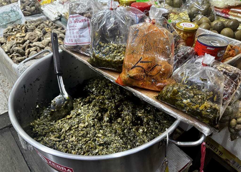 Bangkok Food Tour: Our Honest Review of Chinatown’s Best Street Eats 5 A pot of greens sitting in a giant metal pot is sitting on a shelf in front of other various goods being sold in plastic bags . The shop is in a back street in Bangkok Chinatown.