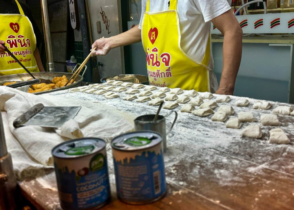 Bangkok Food Tour: Our Honest Review of Chinatown’s Best Street Eats 12 A man in a yellow apron is cutting up dough into pieces to fry into doughnuts at his hawker stall at the Bangkok Chinatown night market.