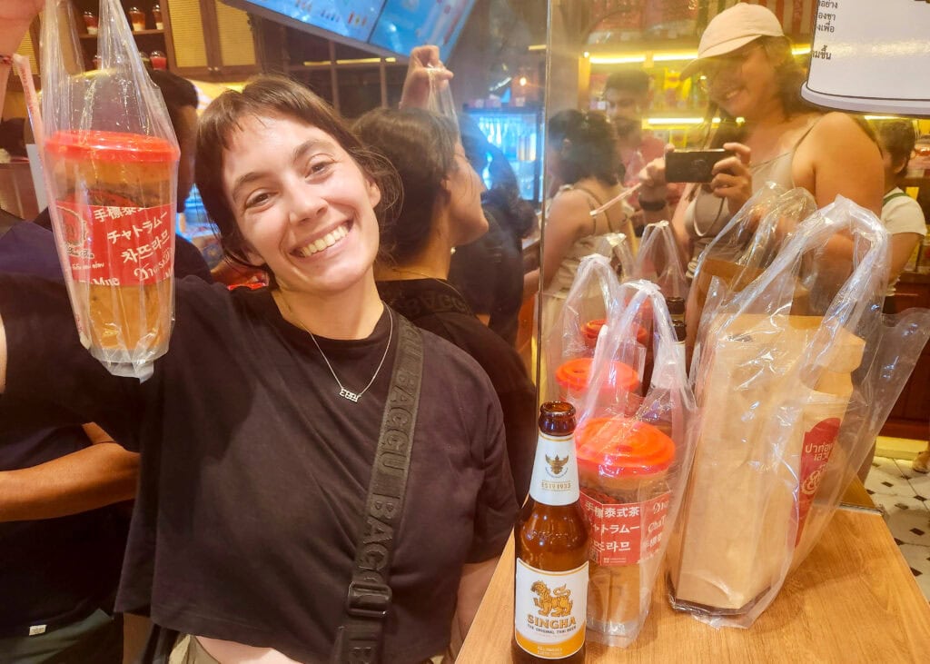 Bangkok Food Tour: Our Honest Review of Chinatown’s Best Street Eats 13 A woman is in a tea shop in Bangkok and is holding up a very large take away thai tea that she was just giving during a Bangkok food tour. There is a half full beer sitting on the table next to her from the previous stop on the tour.