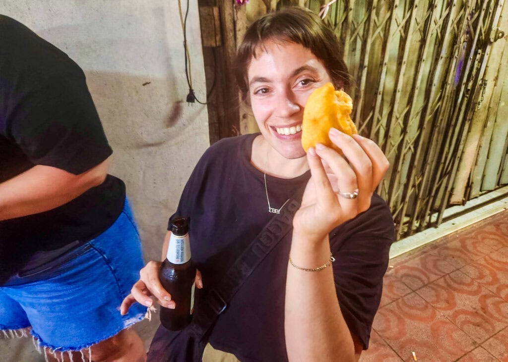 Bangkok Food Tour: Our Honest Review of Chinatown’s Best Street Eats 18 A woman is smiling while standing on the street in Bangkok and holding up a beer and a half eaten donut she was given on a Bangkok food tour.