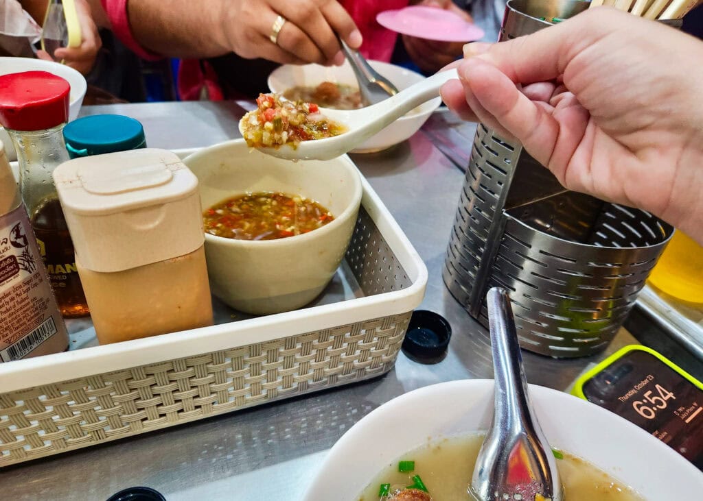 Bangkok Food Tour: Our Honest Review of Chinatown’s Best Street Eats 15 Someone is holding a soup spoon over the metal shop table full of chili vinegar. The sauce is to put in the soup broth that multiple people are eating around the table.
