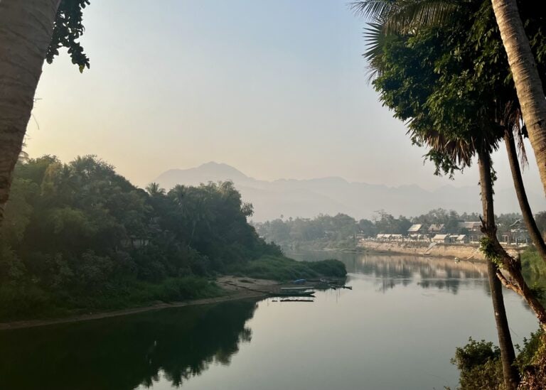 Scenic view of the Nam Khan River at sunrise in Luang Prabang, Laos, with misty mountains and traditional homes in the background."