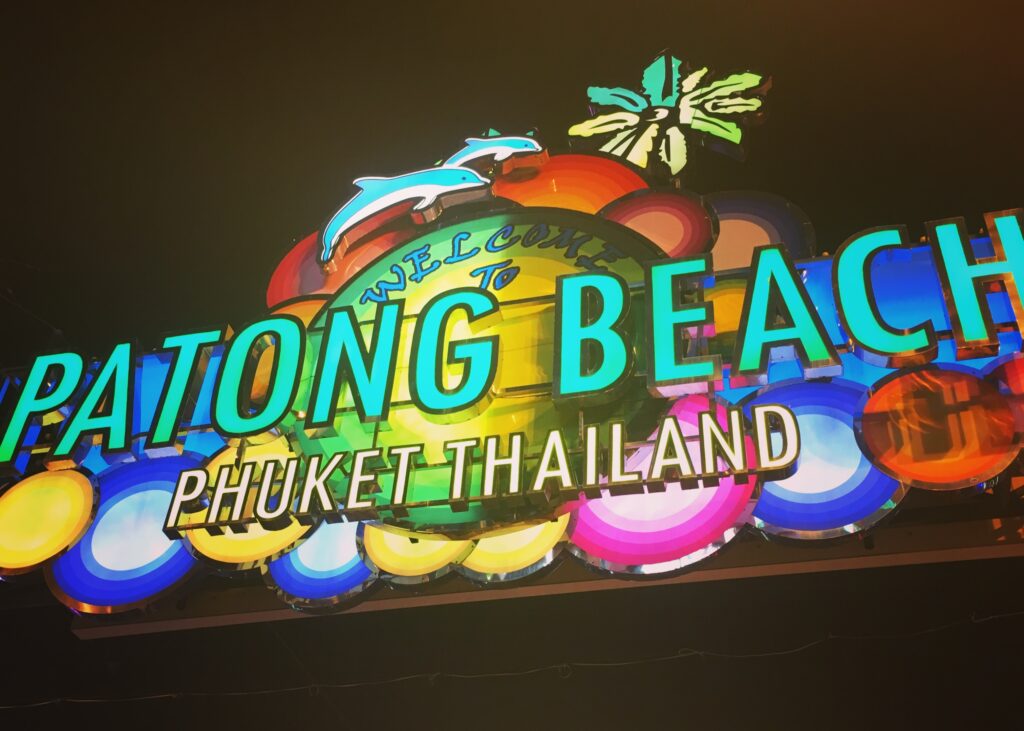 Phuket vs Krabi: Our 20s Party Trip vs 30s Chill Trip 2 The sign to a famous street in Phuket that reads "Patong Beach, Phuket Thailand". It is lit up in neon colours with waves and palm trees behind the text
