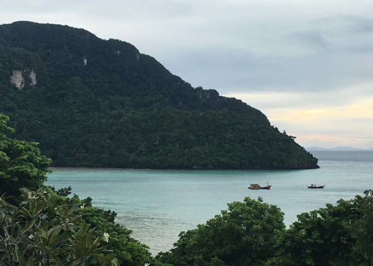 A view of the beach in Thailand with ocean and mountains in the background. There are two boats on the water. A typical view to help decide where is better Phuket vs Krabi.