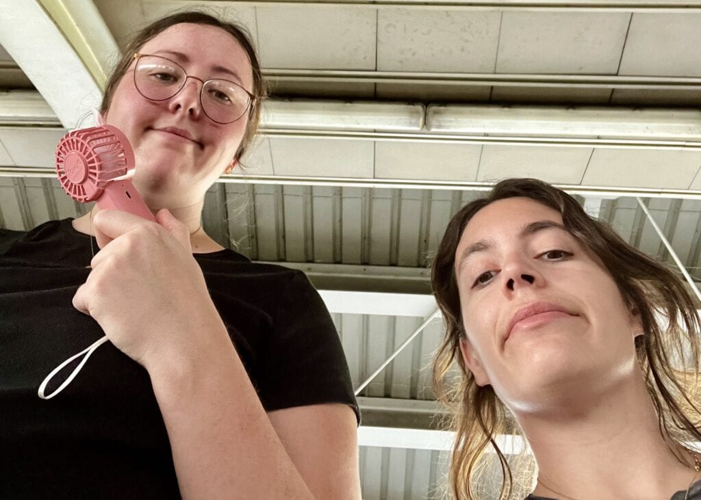 Where to Go in Thailand for the First Time: A Budget Traveler’s Guide 2 Two girls smiling sarcastically at the camera below whilst standing in a train station. They are both sweating and the girl on the left is holding a pink portable fan to her face