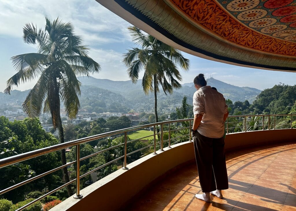 Sri Lanka 2 Traveler standing on a curved balcony overlooking Kandy with palm trees, rolling hills, and a temple roof visible in the distance in Sri Lanka.