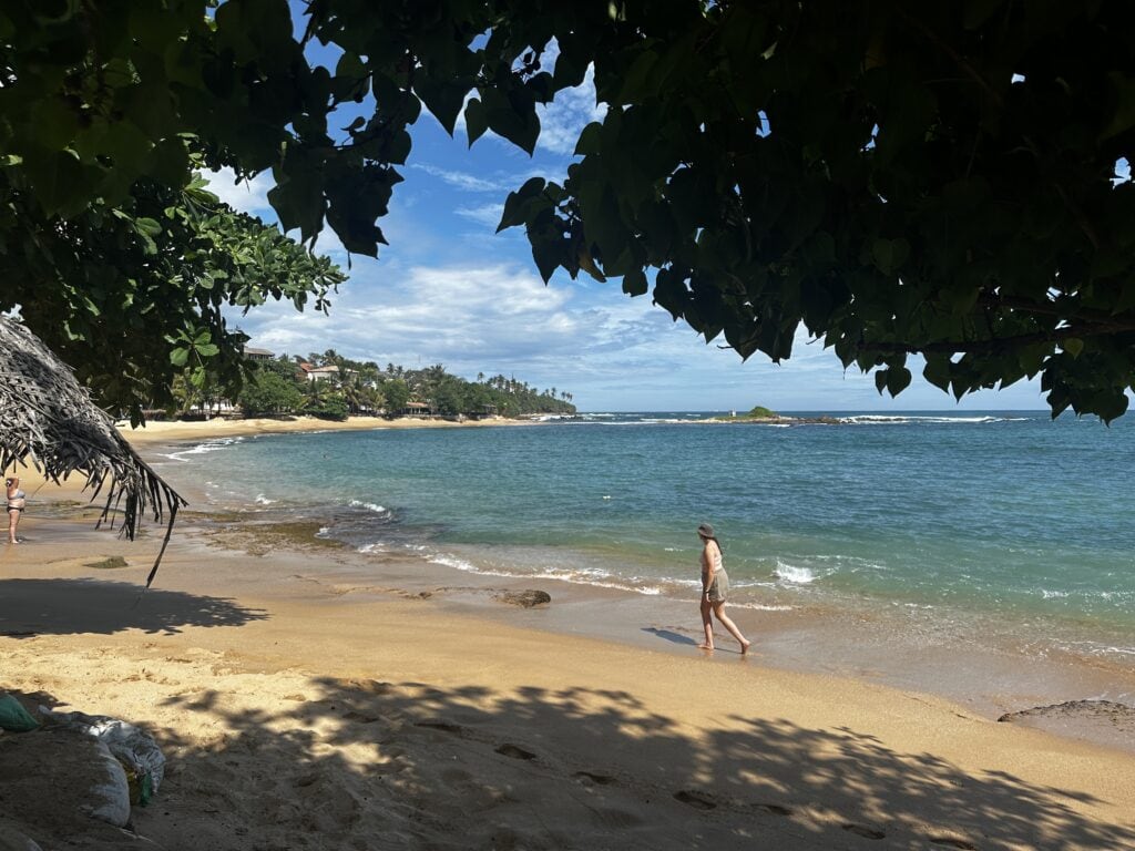 Sri Lanka 1 Quiet tropical beach in Sri Lanka with turquoise water, golden sand, and leafy branches framing the view while a person walks along the shoreline.