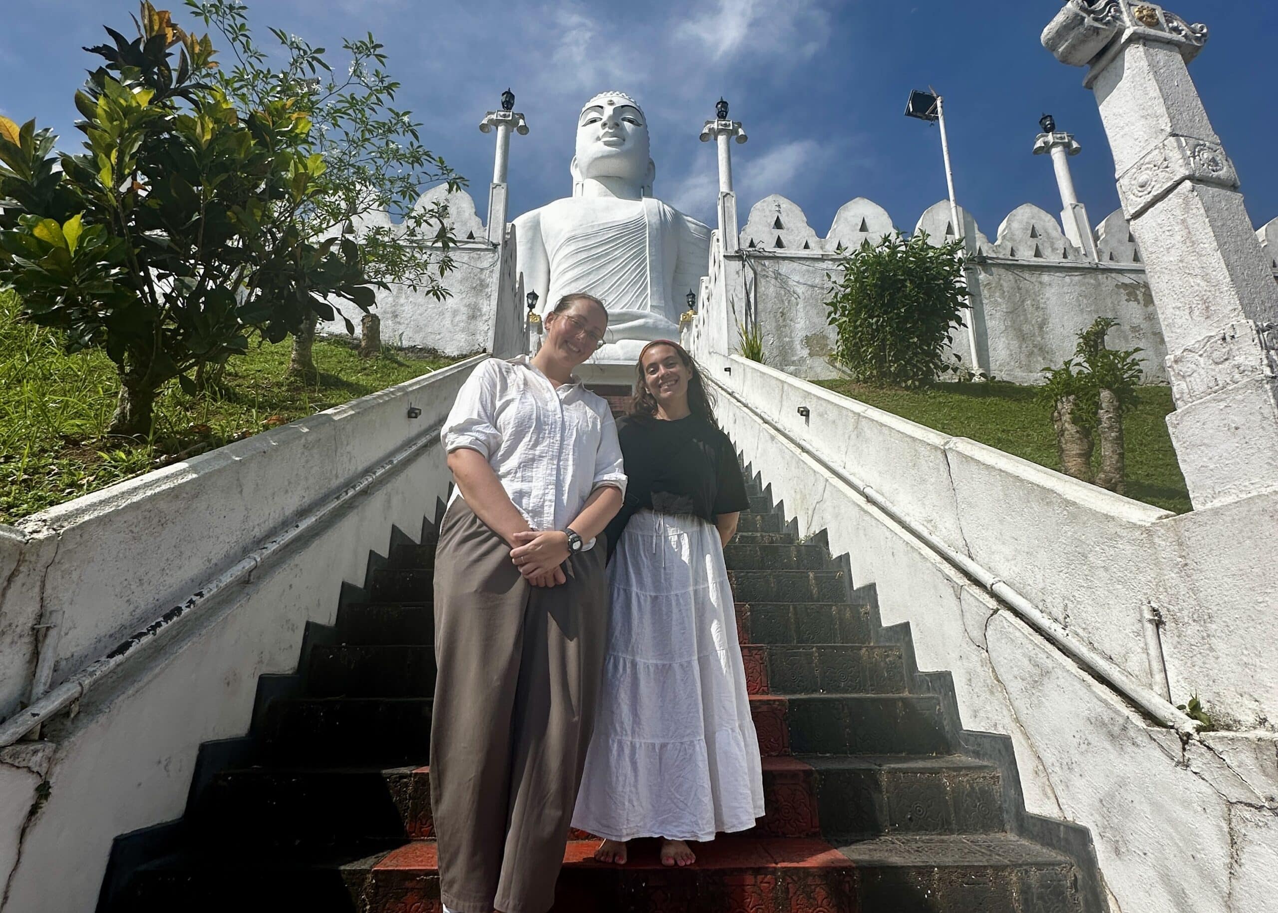 About The Lavender Travelers 5 Two travelers standing on steps leading up to a large white Buddha statue at a temple in Sri Lanka under a blue sky.